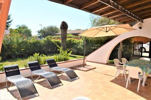 Une terrasse extérieure avec des fauteuils et une table, entourée de verdure et dotée d’un sol carrelé.