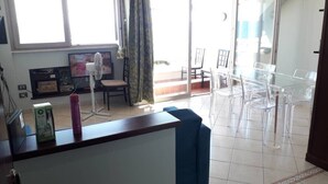 A living room with a sofa, a TV, a fan, and a table with chairs. There is a sliding glass door leading to a balcony with chairs.