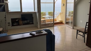 A living room with a TV, a sofa, and a balcony with a view of the sea.