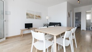 Un comedor moderno con una mesa de madera y sillas blancas, un televisor sobre una mesita auxiliar y una obra de arte en la pared.