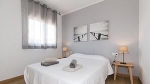 Una cama bien hecha con un edredón blanco, dos mesitas de noche con lámparas y una ventana con cortinas.