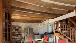 Un intérieur rustique avec une table à manger en bois, des chaises rouges, un sol en pierre, un coin cuisine et un escalier en bois.