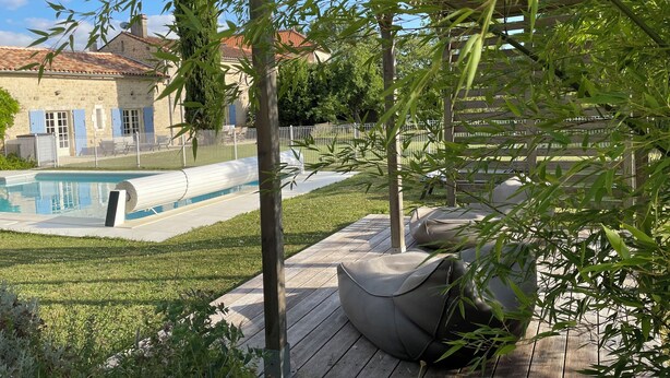 Un espace piscine doté d’une terrasse en bois, de sièges extérieurs et offrant une vue sur un bâtiment en pierre aux volets bleus.