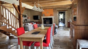 Un intérieur rustique avec une table à manger en bois, des chaises à l’assise rembourrée de couleur rouge, une cheminée en pierre et un escalier.