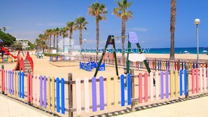 Een strand met een houten omheining, palmbomen en speeltoestellen.