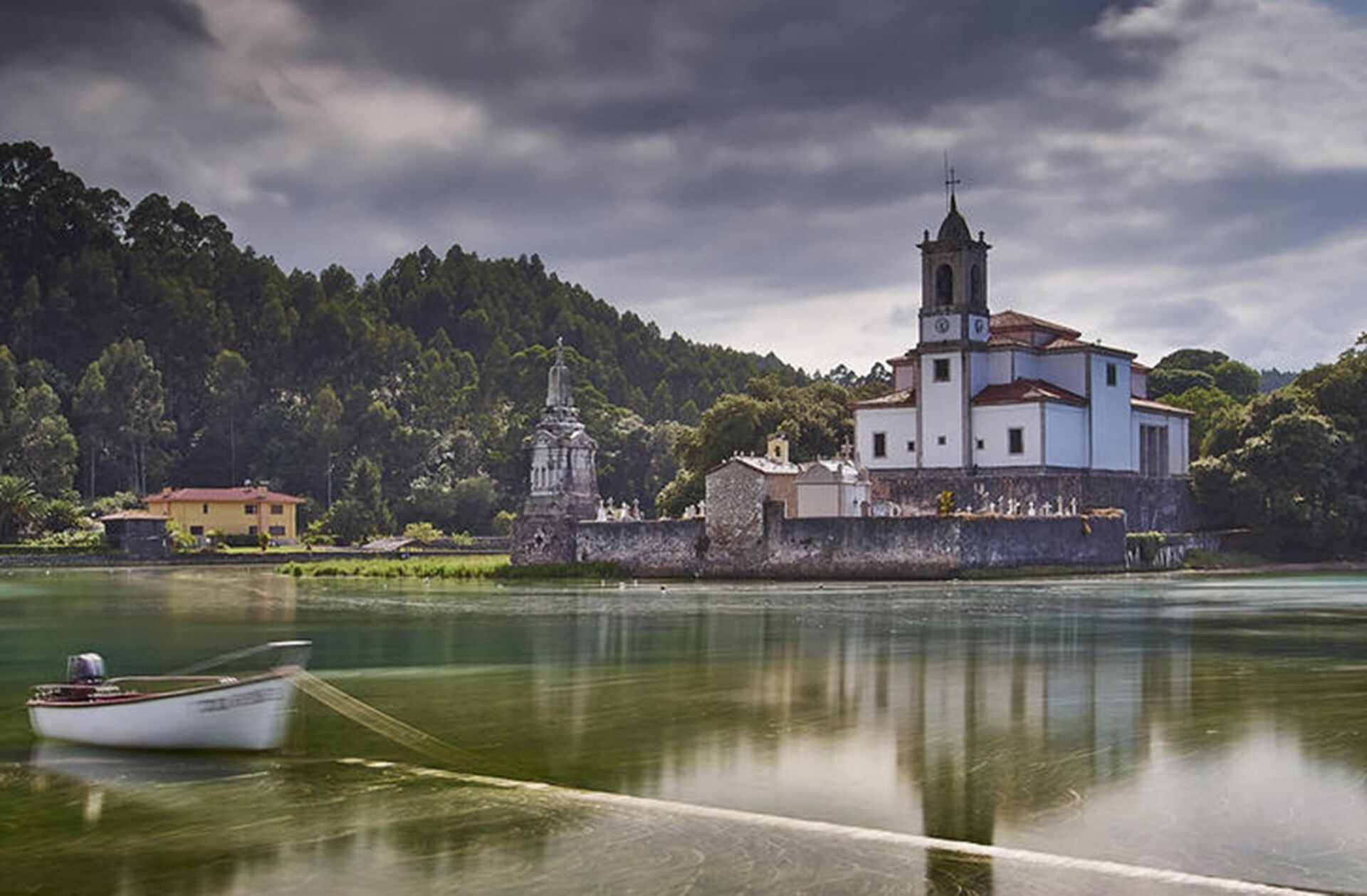 A white church with a bell tower by a calm lake, surrounded by greenery and a small boat anchored nearby.
