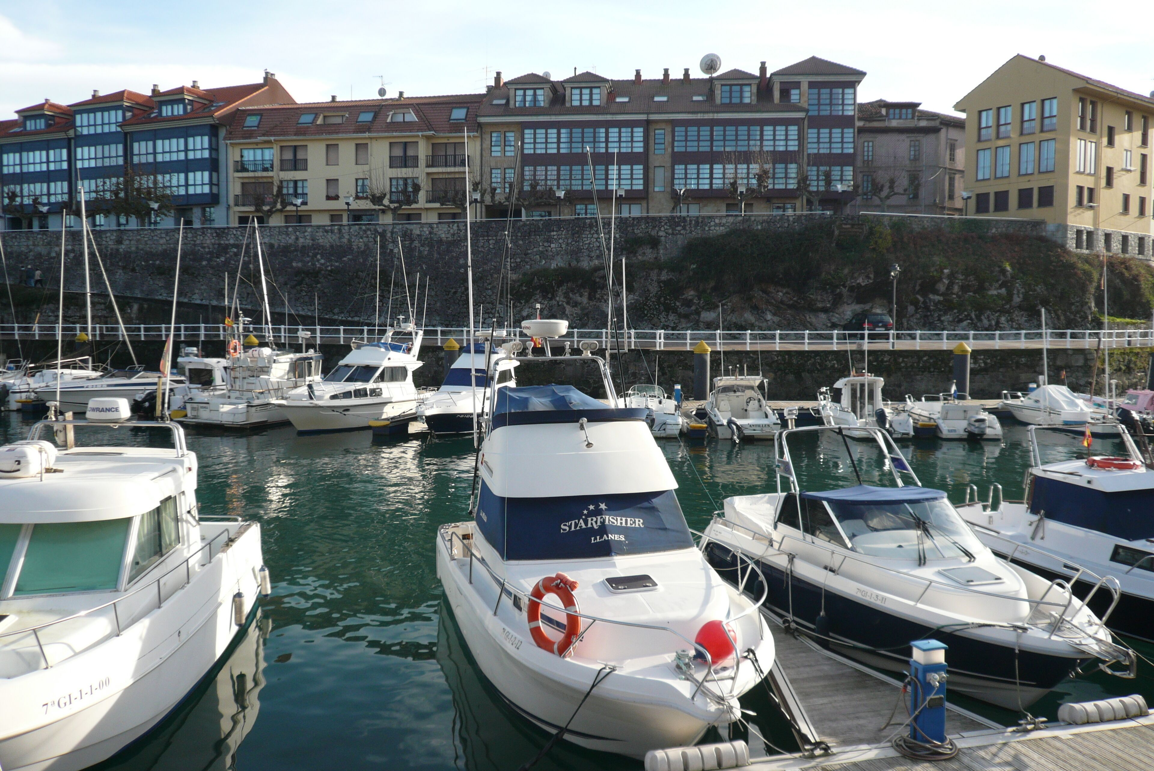 Un port de plaisance où sont amarrés de nombreux bateaux, un mur de pierre et des bâtiments en arrière-plan.