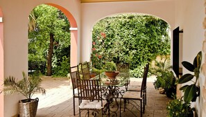 Um pátio com mesa e cadeiras, cercado por plantas em vasos e um fundo verde exuberante.