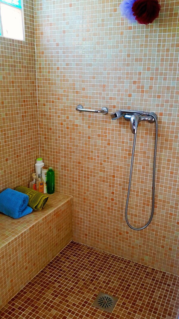 A shower area with a tiled wall, a handheld showerhead, and a drain. There are towels and toiletries on a bench.