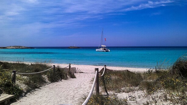 Een zandstrand met een touwbarrière en een helderblauwe zee. Een zeilboot is in de verte te zien.