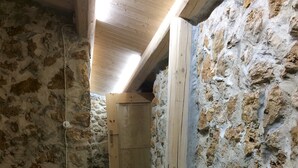 A stone-walled room with a wooden bench, a wicker basket, and a door leading to another room. - House Mmiriusa (Noto)