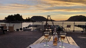 Ett träbord med en duk med prickmönster, två ölglas och en flaska sprit står på en brygga med utsikt över en vattenyta. I bakgrunden syns båtar och kullar.