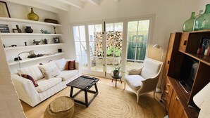 Ein Wohnzimmer mit einem weißen Sofa, einem Holztisch, einem Beistelltisch mit einer Pflanze, einem Sessel und einem Bücherregal mit grünen Flaschen.