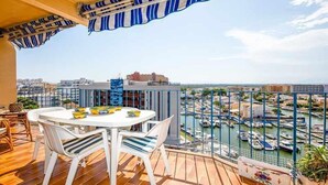 Un balcon avec une table et des chaises blanches, un auvent rayé et une vue sur une marina remplie de bateaux.