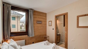 A bedroom with a wooden wall, a large window with curtains, a bed with white bedding, and a small bedside table. - Apartment Staddon - Alpes Agency - Central Chamonix - Sleeps 6 (Chamonix-Mont-Blanc)