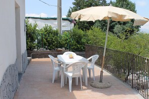 Een terras buiten met een tafel en stoelen, een beige parasol en uitzicht op bomen en bergen.