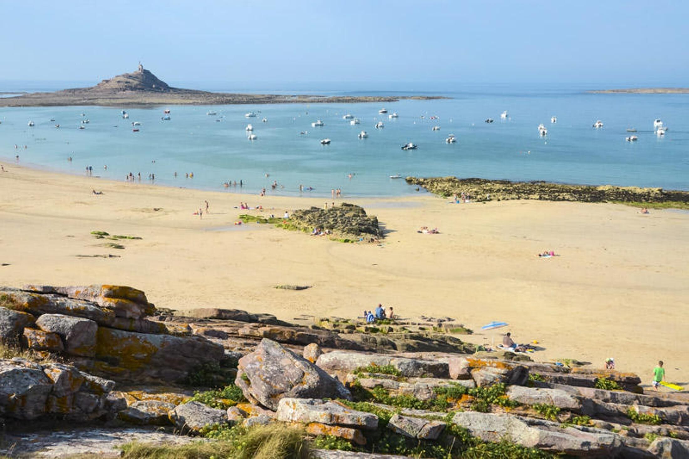 A beach with clear skies, rocky foreground, and calm sea with boats.