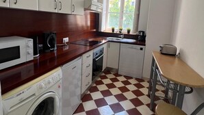 Une cuisine compacte avec des meubles blancs, un sol à carreaux rouges et blancs, un lave-linge, une machine à café et une petite table avec une chaise.