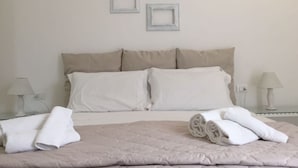 Un lit bien fait, recouvert d’une courtepointe beige, avec deux oreillers blancs et des serviettes blanches roulées posées sur le côté. Trois tableaux encadrés sont accrochés au mur au-dessus du lit.