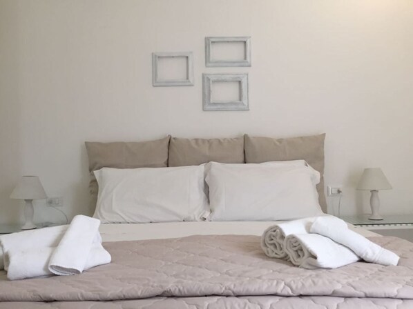 Una cama bien hecha con colcha color beige, dos almohadas blancas y toallas blancas enrolladas al lado. Encima de la cama, hay tres cuadros en la pared.