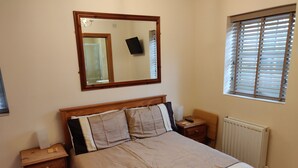Une chambre à coucher comprenant un lit, une table de chevet, une commode, un miroir, une fenêtre avec des stores et un radiateur.