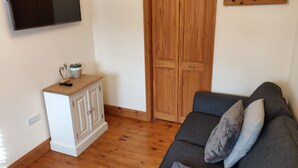 Un salon avec un plancher en bois, une armoire blanche, un canapé gris avec des coussins, une porte en bois et un téléviseur fixé au mur.