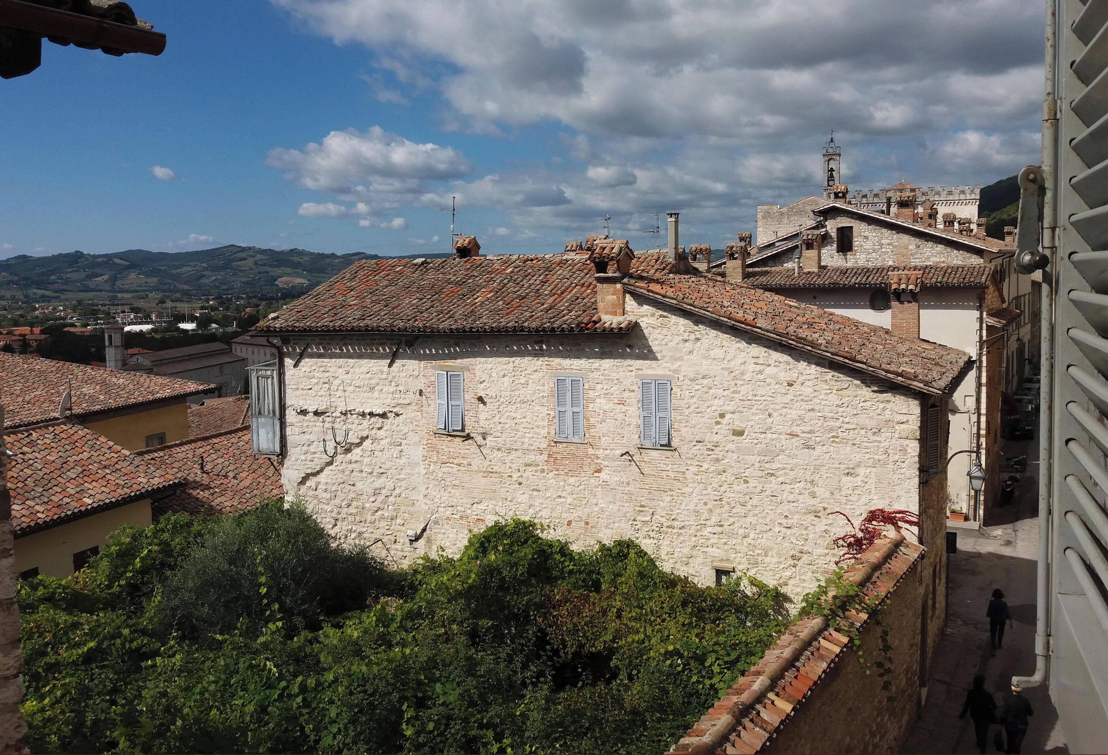 A stone building with a tiled roof and shutters, surrounded by greenery and other buildings.