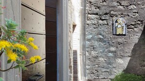 Un mur de pierre orné d’une icône religieuse, une porte en bois et une plante aux fleurs jaunes.