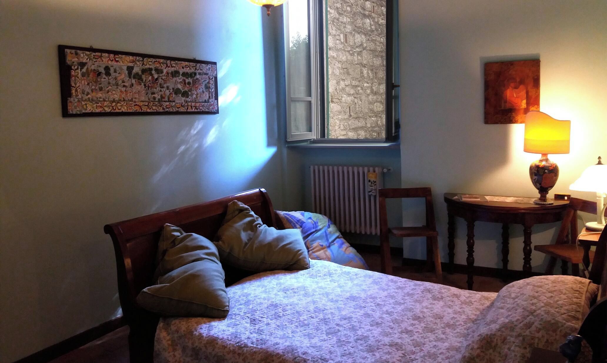 A bedroom with a bed, a chair, a table, a lamp, a radiator, and a window.