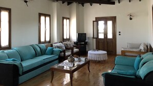 A spacious living room with a high ceiling, wooden beams, and large windows. There are two sofas, a coffee table, and a television. - Villa Maira, Chalkidiki. Holidays as it should be! (Afytos Kassandras Chalkidiki)