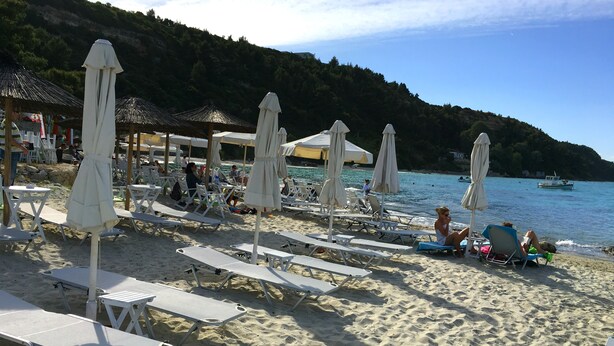 A beach with rows of white lounge chairs and umbrellas, a calm sea, and a hilly backdrop. - Villa Maira, Chalkidiki. Holidays as it should be! (Afytos Kassandras Chalkidiki)