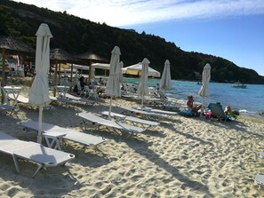 A beach with rows of white lounge chairs and umbrellas, a calm sea, and a hilly backdrop. - Villa Maira, Chalkidiki. Holidays as it should be! (Afytos Kassandras Chalkidiki)