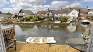 A wooden deck with a boat docked by a calm canal, surrounded by residential houses. - Self Catering 3 Bedroom Sunset  Haven In Wroxham, Norfolk, Norfolk Broads, Eng (Wroxham)