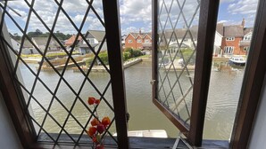 A view through a window with a diamond-patterned grille, overlooking a calm river with boats and houses in the background. - Self Catering 3 Bedroom Sunset  Haven In Wroxham, Norfolk, Norfolk Broads, Eng (Wroxham)