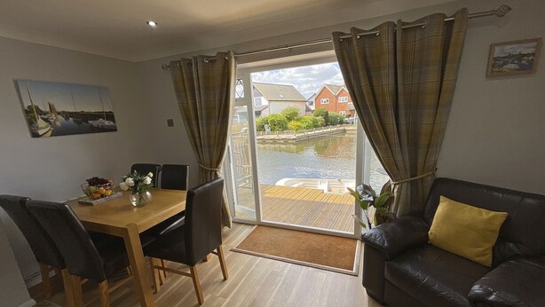 A room with a dining table, chairs, a sofa, and a view of a water body outside. - Self Catering 3 Bedroom Sunset  Haven In Wroxham, Norfolk, Norfolk Broads, Eng (Wroxham)