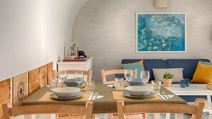 Ein Essbereich mit einem Tisch für vier Personen, Holzstühlen, einem blauen Sofa mit Kissen und einer Wandlampe.