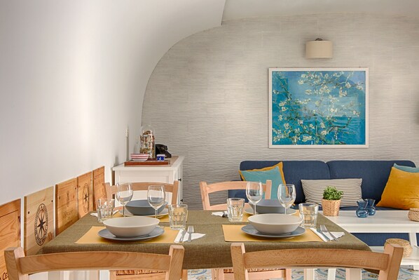 A dining area with a table set for four, wooden chairs, a blue sofa with cushions, and a wall-mounted lamp.