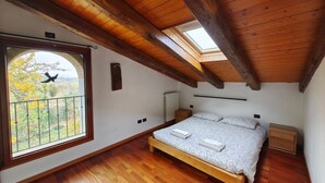 Quarto com teto de madeira, claraboia, cama com almofadas, mesa de cabeceira e janela com vista para árvores.