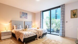 Ein modernes Schlafzimmer mit zwei Betten, einem großen Fenster mit Vorhängen, einem Blumentopf und einem Wandbild.