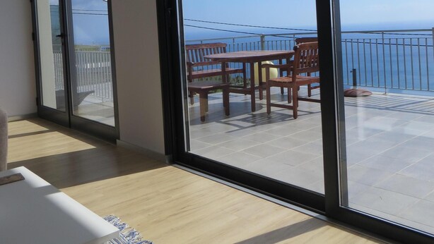 Ein geräumiger Balkon mit einem Holztisch und Stühlen, großen Fenstern und Blick aufs Meer.