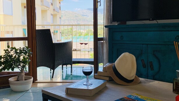 Una habitación con vistas a un balcón, una mesa con un libro, una copa de vino, un sombrero y una revista.