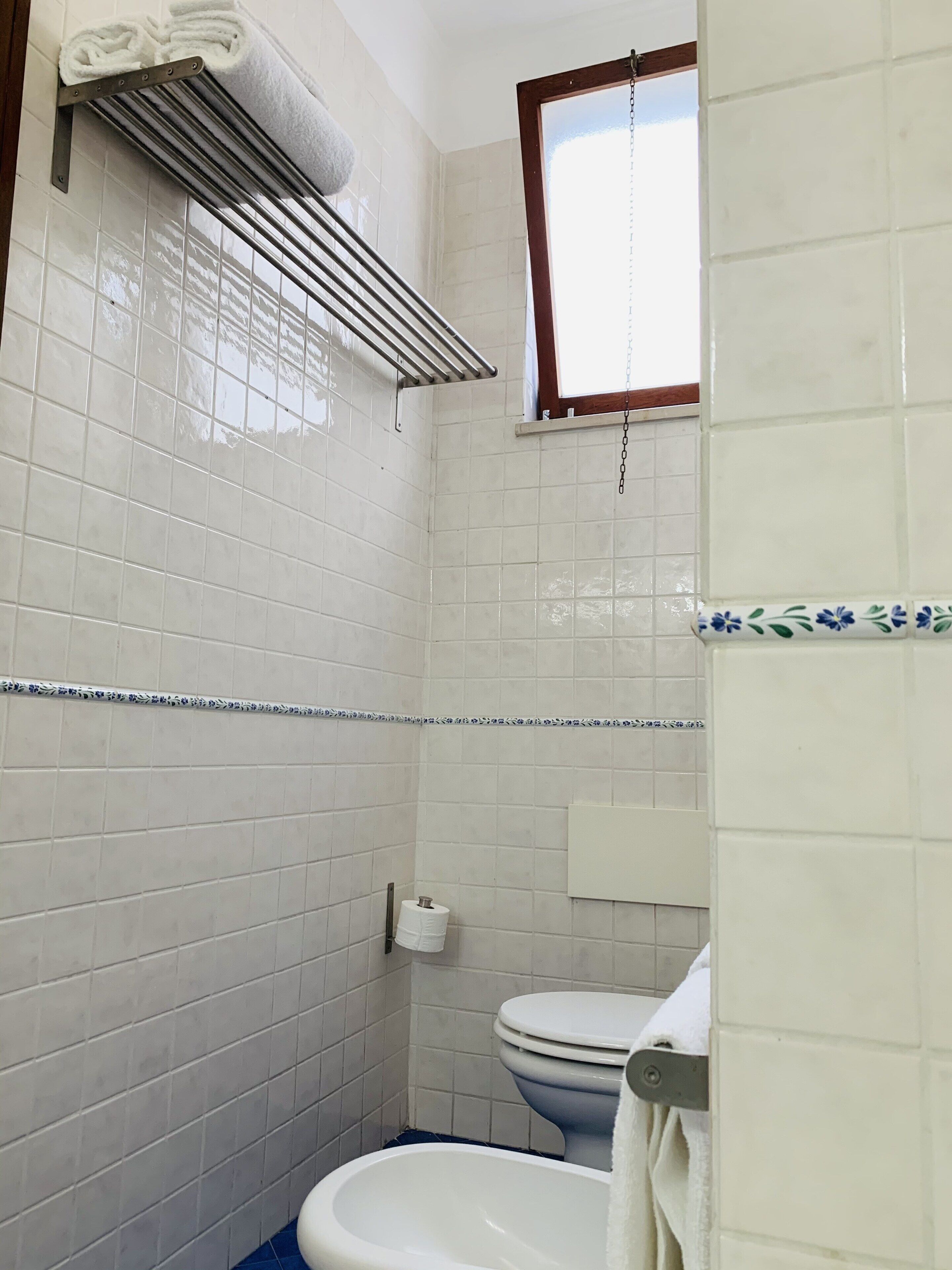 A bathroom with white tiled walls, a toilet, a towel rack, and a window.