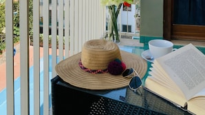 Un balcón con vistas a una colina, una mesa con un sombrero, gafas de sol y un libro abierto, y un jarrón con flores.