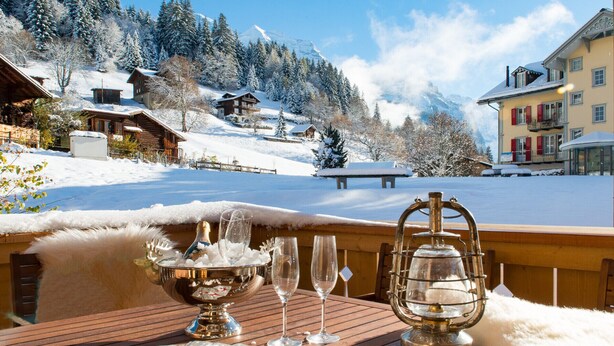 A wooden deck with a table set for a meal, overlooking a snowy mountain landscape with buildings. - Panoramapark Soleil Camelia Apt. Akroyd (Wengen)