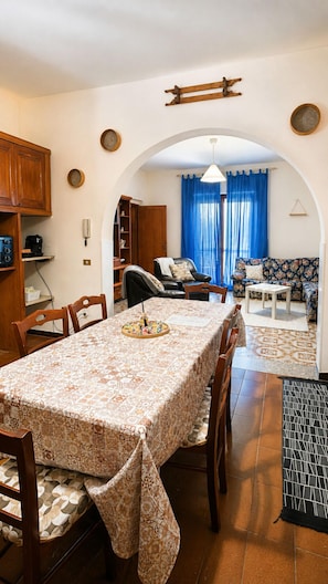 Un comedor con mesa, sillas y una zona de cocina con armarios y una cafetera.