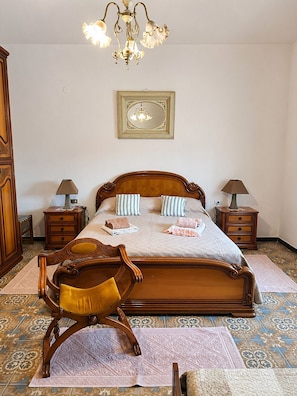 Un dormitorio con una cama de madera grande, dos mesitas de noche, una silla de madera, una araña y un cuadro enmarcado en la pared.