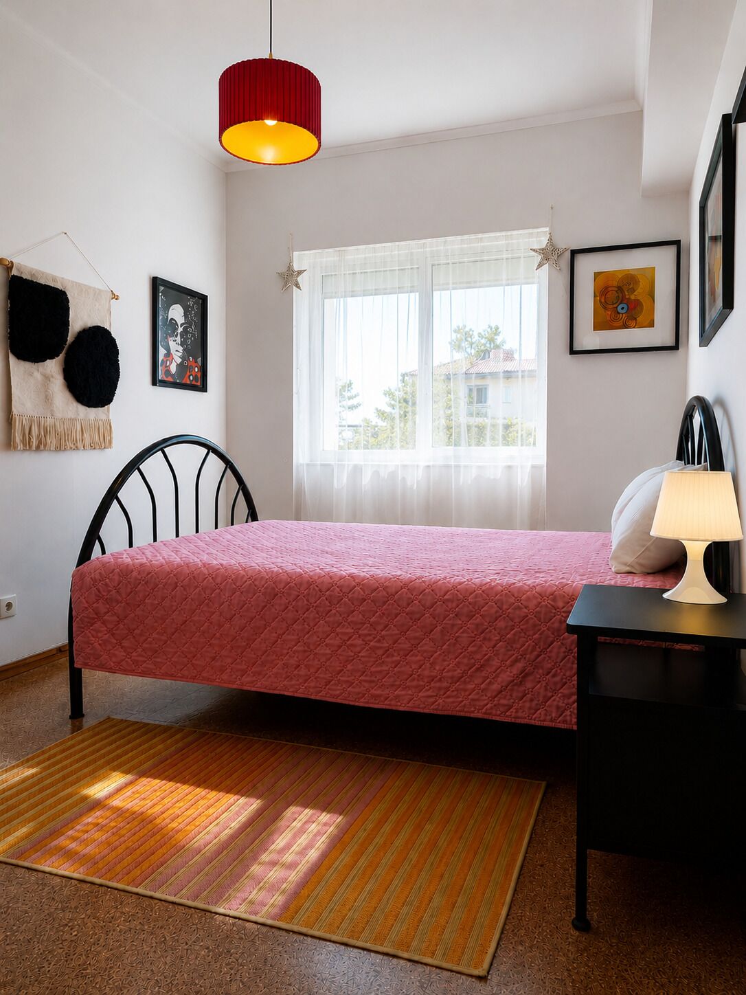 Une chambre à coucher avec un lit, une table de chevet, une lampe, une fenêtre avec des rideaux, une décoration en forme d’étoile et un tableau encadré accroché au mur.
