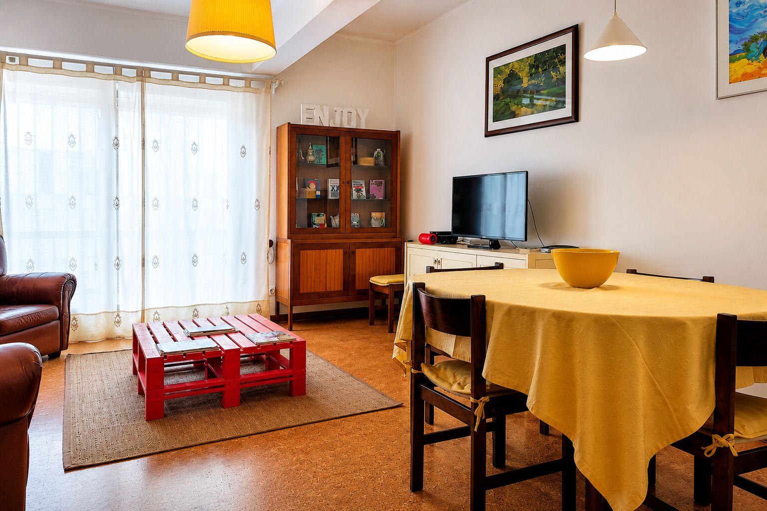 Een woonkamer met een bank, een houten kast, een televisie en een eettafel met een gele tafelkleed.