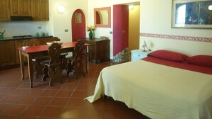 A bedroom with a bed, a desk, and a chair. There is a kitchen area with cabinets and a stove.