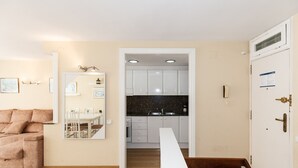 Una sala moderna con un sofá color beige, un comedor con una mesa y sillas blancas, y una cocina con gabinetes blancos y una encimera de granito.